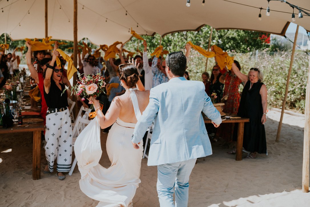 The couple walking into the stretch tent on the sand while guests wave their napkins